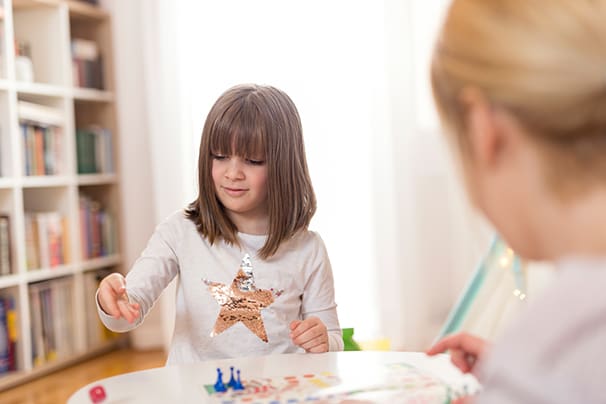 Mother and daughter sitting in a playroom, playing a ludo game; daughter rolling the dice